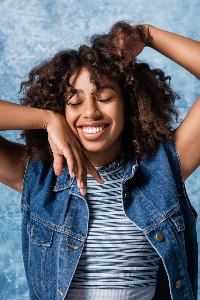 stylish african american woman with closed eyes touching hair while holding hand near face on blue background - Fotó, kép
