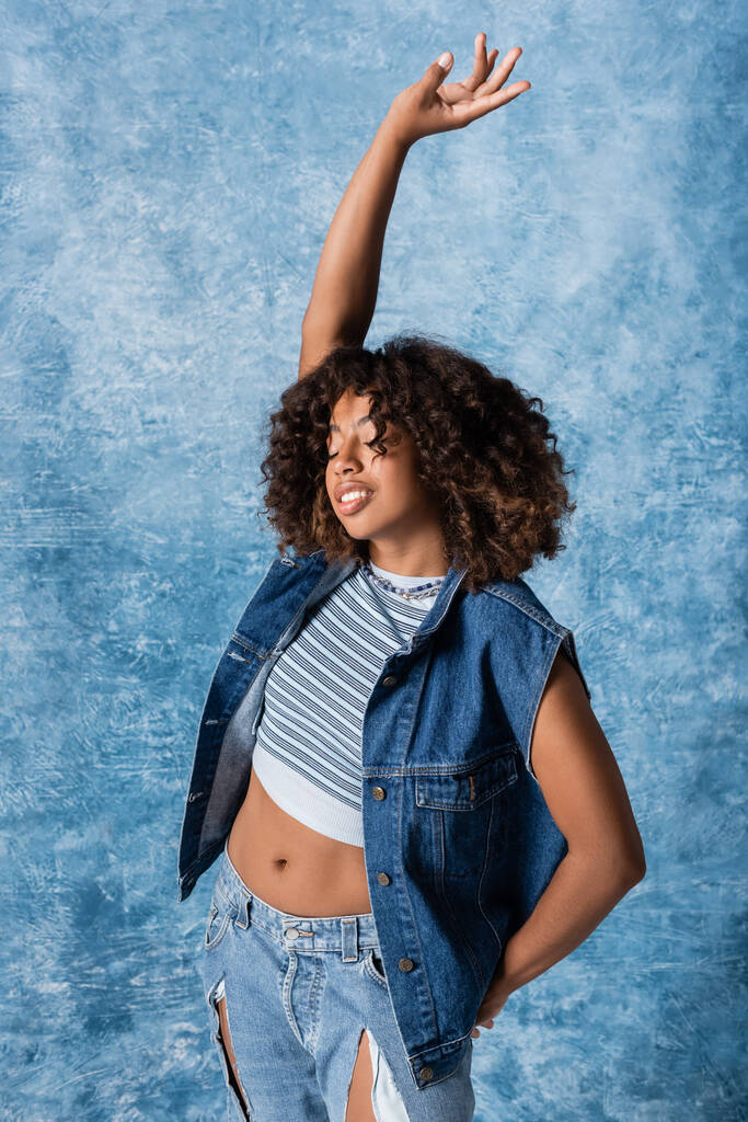 african american woman in denim vest and crop top standing with closed eyes and raised hand on blue background - 写真・画像
