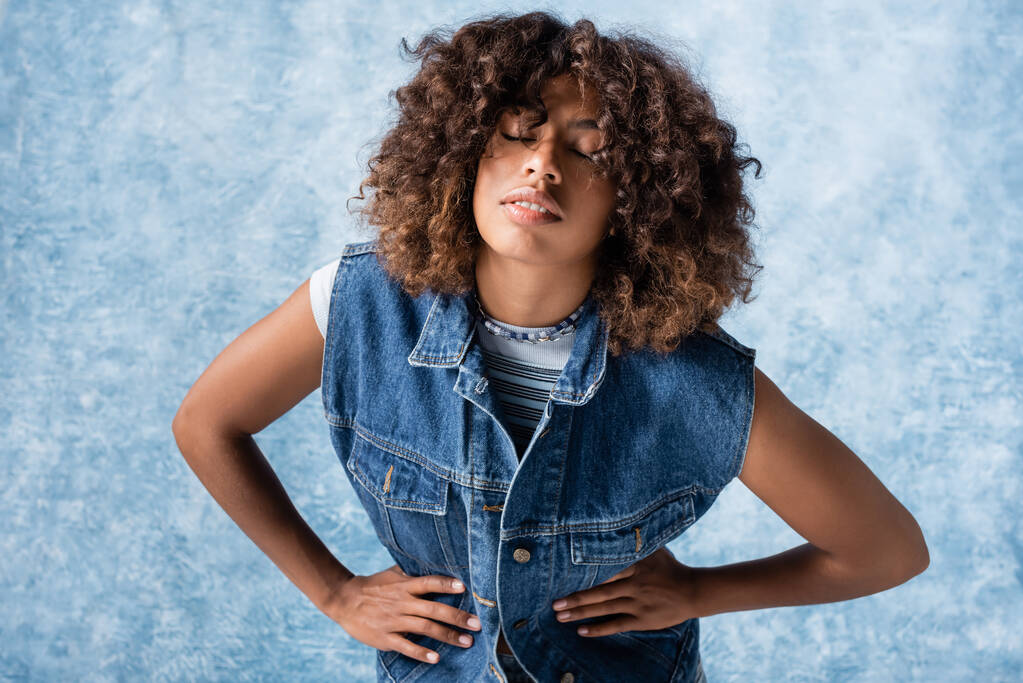 stylish african american woman in denim vest standing with hands on waist on blue background - Zdjęcie, obraz