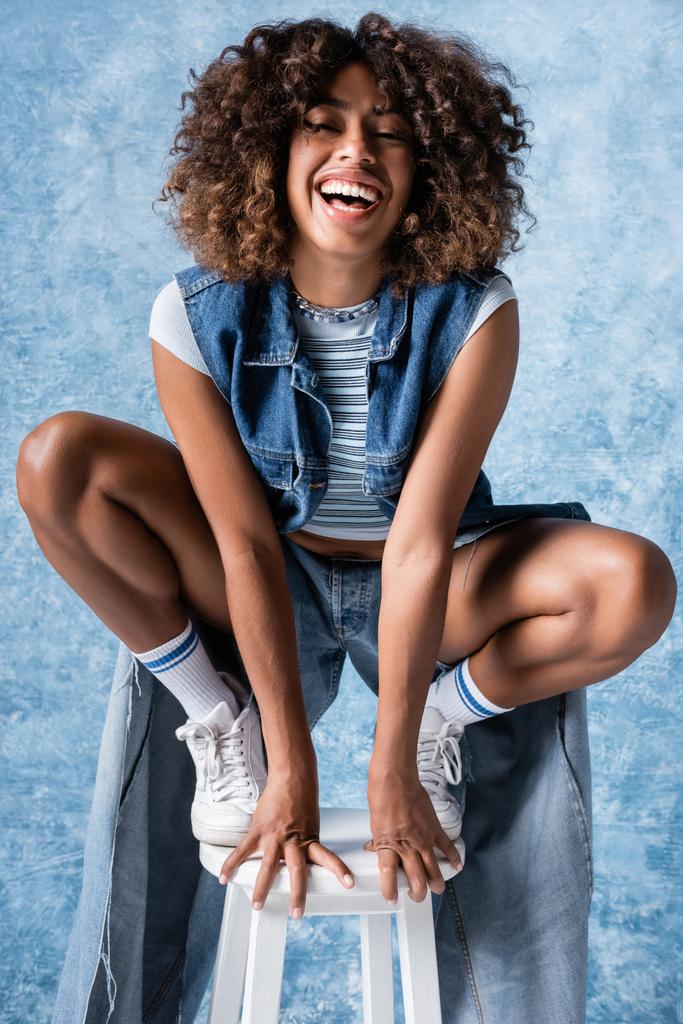 laughing african american woman in denim clothing posing on haunches on blue background - Foto, immagini