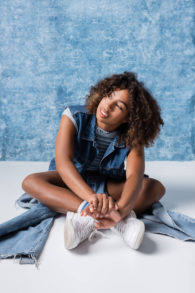 african american woman in white sneakers and denim clothes sitting with crossed legs on blue background - Фото, зображення
