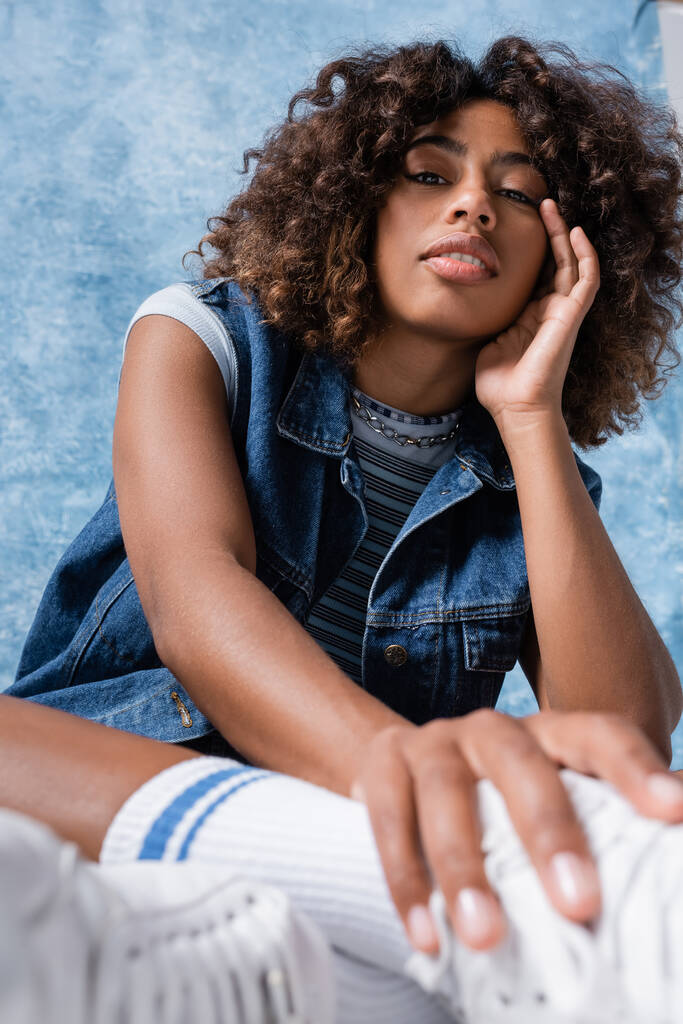 trendy african american woman in denim vest holding hand near face and looking at camera on blue background - Fotó, kép
