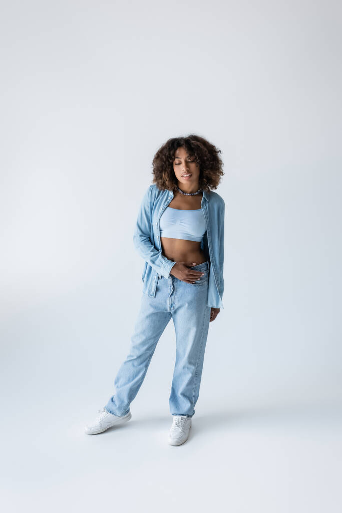full length of african american woman in white sneakers and blue denim clothing on grey background - Foto, imagen