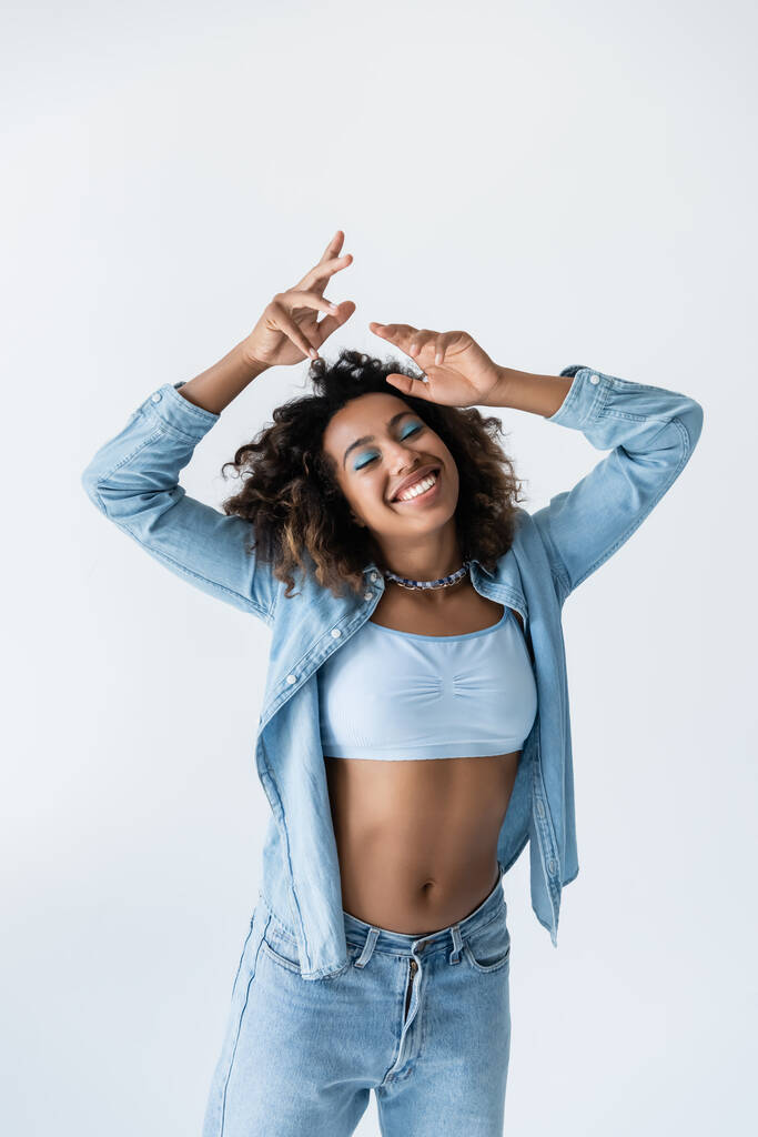 усміхнена і модна афроамериканка з закритими очима стоїть руками над головою ізольована на сірому
 - Фото, зображення