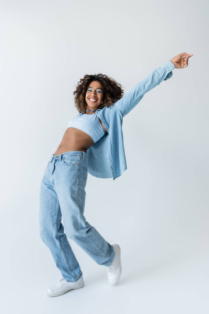 full length of excited african american woman in crop top and blue denim clothes on grey background - Фото, изображение