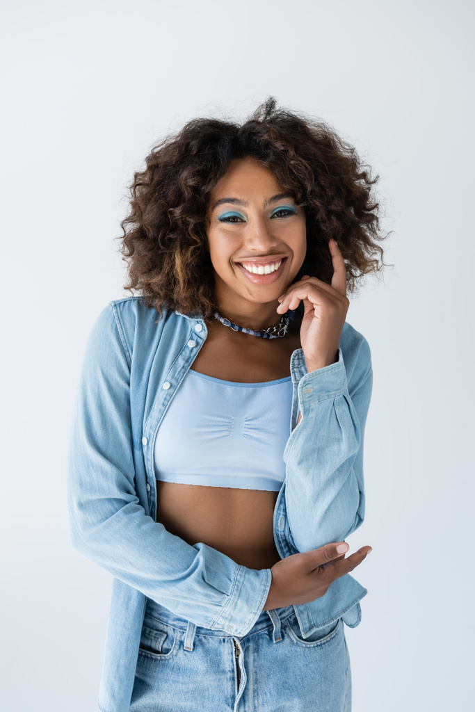 positive african american woman in white crop top and denim shirt looking at camera isolated on grey - Photo, image
