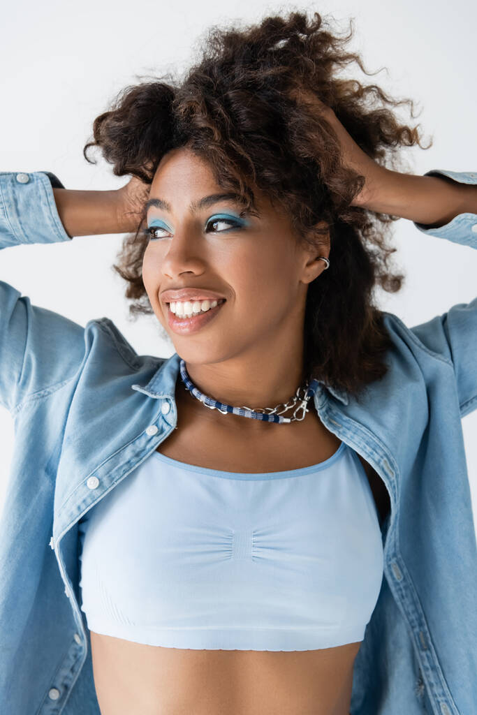 smiling african american woman in necklaces and denim shirt touching wavy hair isolated on grey - Fotoğraf, Görsel