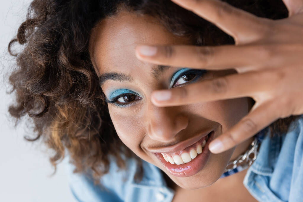 cheerful african american woman with makeup obscuring face with hand isolated on blue - 写真・画像