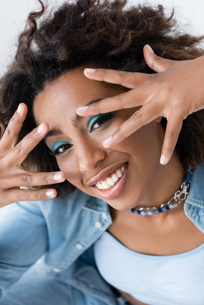 щаслива афроамериканка затемнює обличчя руками, дивлячись на камеру ізольовано на сірому
 - Фото, зображення