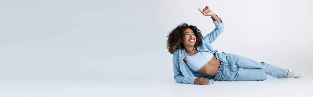 щаслива афроамериканська жінка в джинсовому одязі, що лежить і вказує пальцем на сірому фоні, банер
 - Фото, зображення