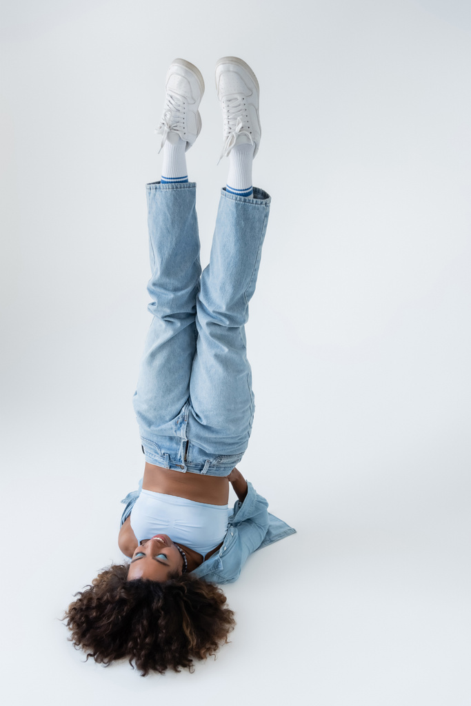 african american woman in blue jeans and white sneakers in shoulder stand pose on grey background - Photo, image