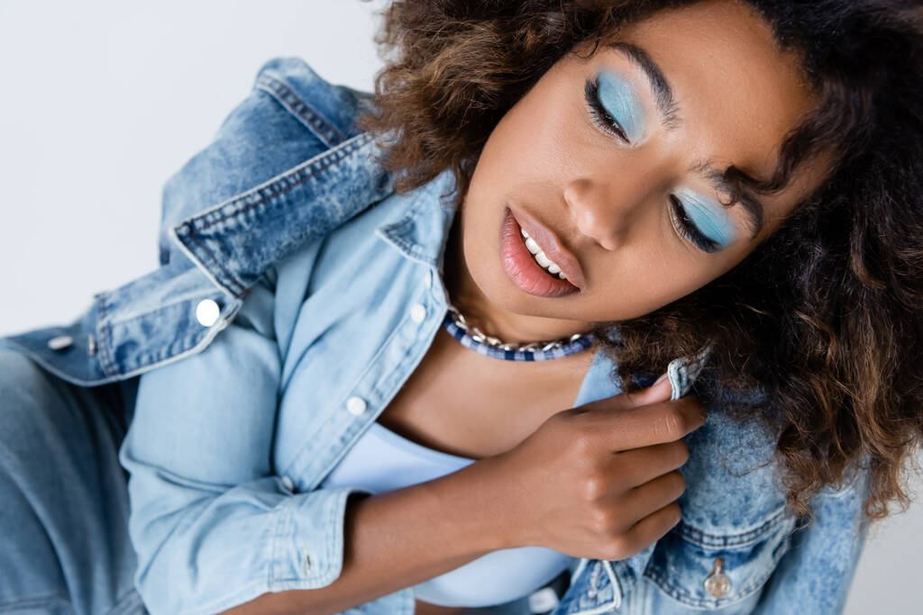 sensual african american woman with blue eye shadows posing with denim garments isolated on grey - Foto, immagini