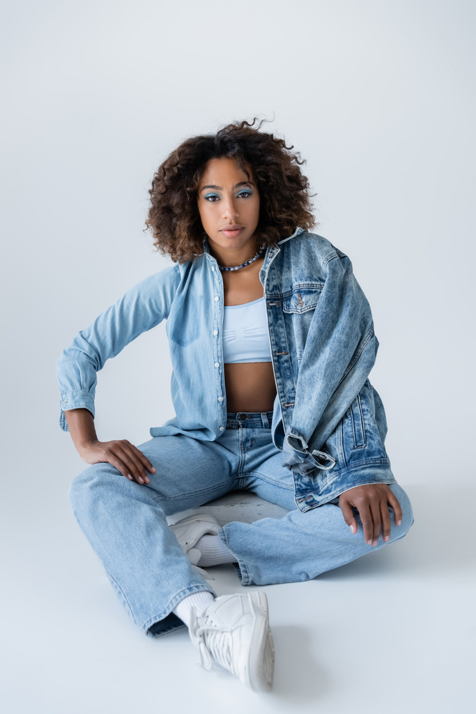 curly african american woman in blue jeans and sneakers sitting on grey background - Foto, imagen