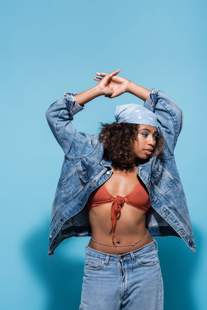fashionable african american woman in bandana and denim jacket standing with hands above head on blue background - Foto, afbeelding