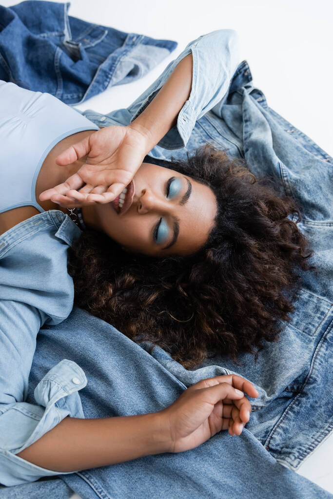 top view of african american woman lying with hand near face near denim clothing on grey background - Foto, Bild