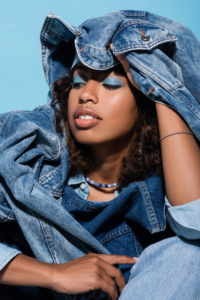 sensual african american woman covering head with denim jacket isolated on blue - Foto, Imagem