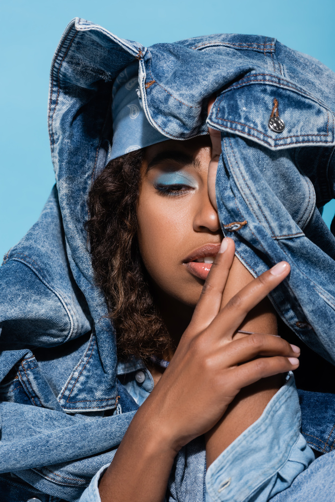 african american woman with makeup obscuring face with denim jacket isolated on blue - Zdjęcie, obraz