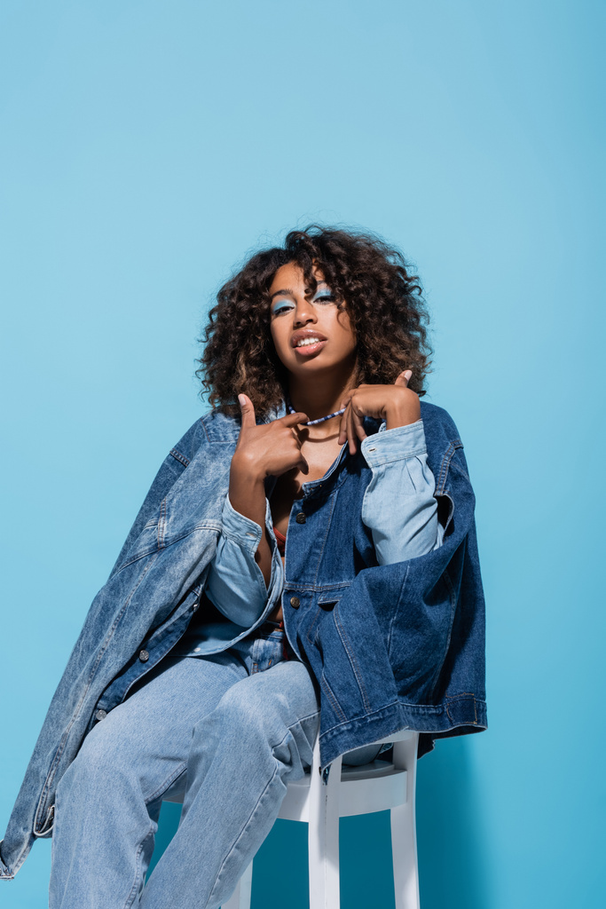 african american woman in denim clothes touching necklace while sitting on blue background - Fotó, kép