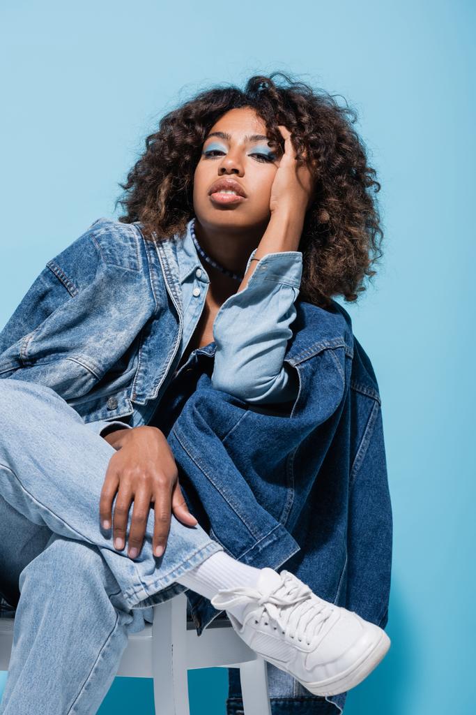 african american woman in trendy denim clothing looking at camera while sitting on blue background - Zdjęcie, obraz