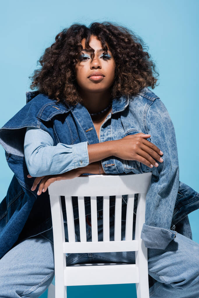 stylish african american woman with wavy hair sitting on chair with crossed arms isolated on blue - Foto, Imagem