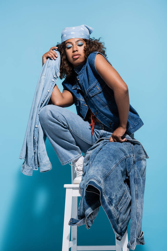 african american woman posing on chair with denim garments on blue background - Valokuva, kuva