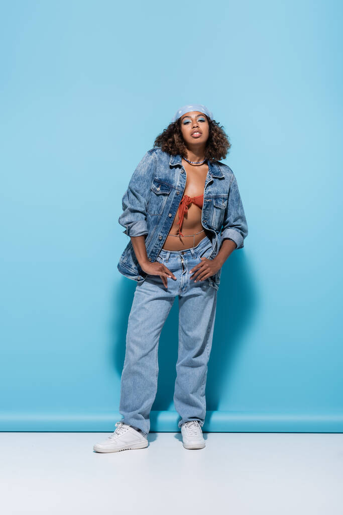 full length of african american woman in denim clothes posing with thumbs in pockets on blue background - Foto, afbeelding