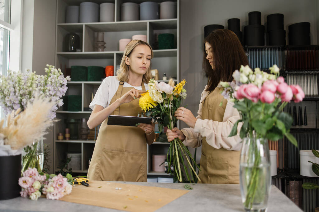 Portrait of two female florists decorating bouquet, watching instruction on tablet working in flower shop. Woman sales assistant working as florist and holding bouquet with business owner together - Foto, immagini