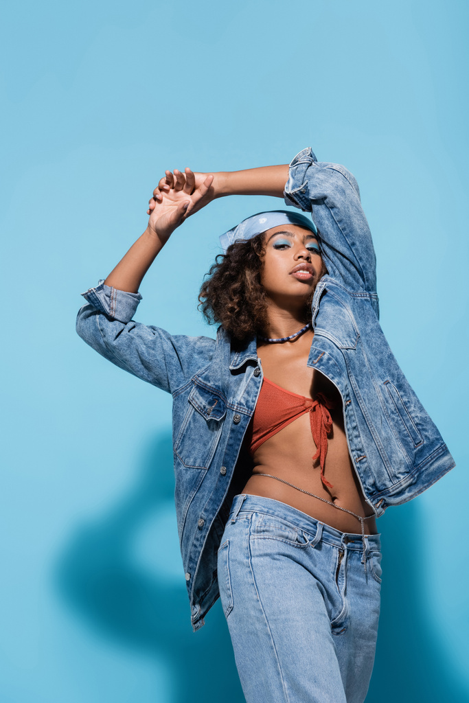 fashionable african american woman in denim jacket and bandana standing with hands above head on blue background - 写真・画像
