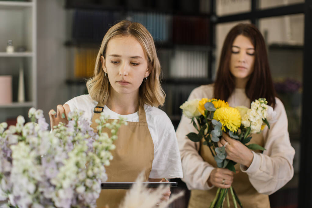 Дві усміхнені жінки-флористи працюють за лічильником в сучасному інтер'єрі лофт квіткового магазину. Флористика, бізнес, концепція прикраси. Творче заняття в квітковому бутіку
. - Фото, зображення