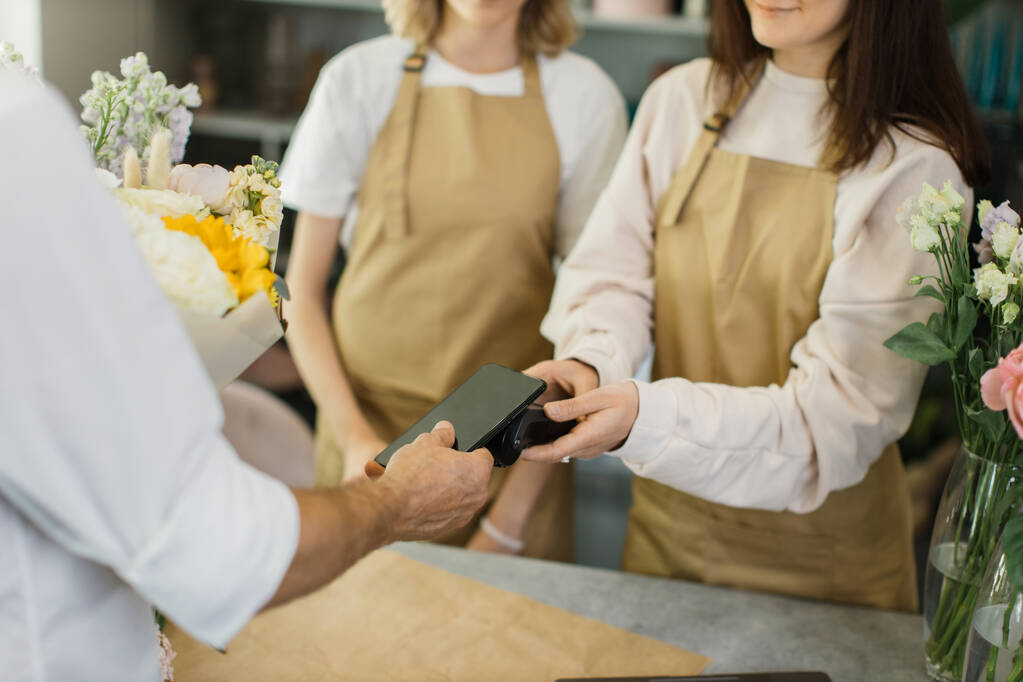 Close up of caucasian male hands using electronic credit card on his smartphone paying for purchase using payment terminal in flower shop. Client buying bouquet making payment in store - Foto, Imagem