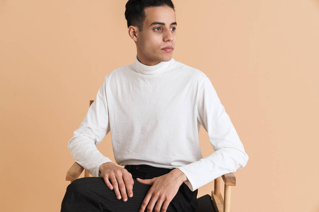 Young middle eastern man looking aside while sitting on chair isolated over beige background - Φωτογραφία, εικόνα