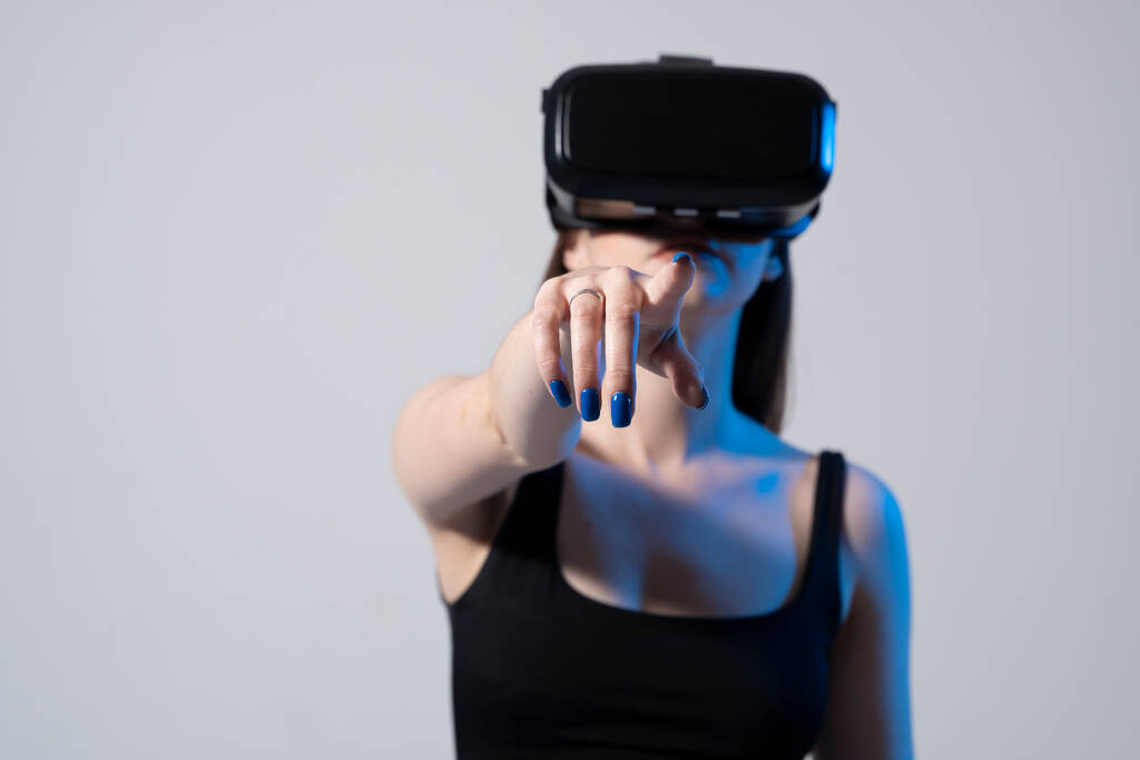 Młoda atrakcyjna kobieta w czarnej bluzce i kasku VR stoi i dotyka powietrza. Pojęcie nowoczesnej technologii i wirtualnej rzeczywistości - Zdjęcie, obraz