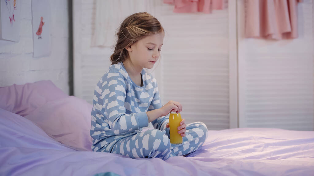 Preteen child in pajama holding bottle with orange juice on bed  - 写真・画像