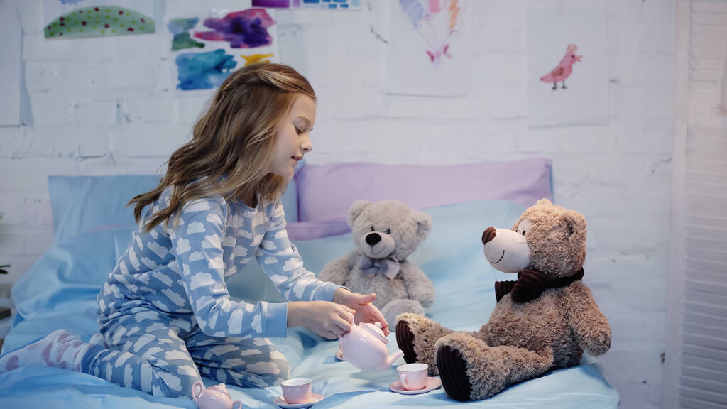 Side view of smiling preteen kid pouring tea near soft toys on bed  - Fotoğraf, Görsel
