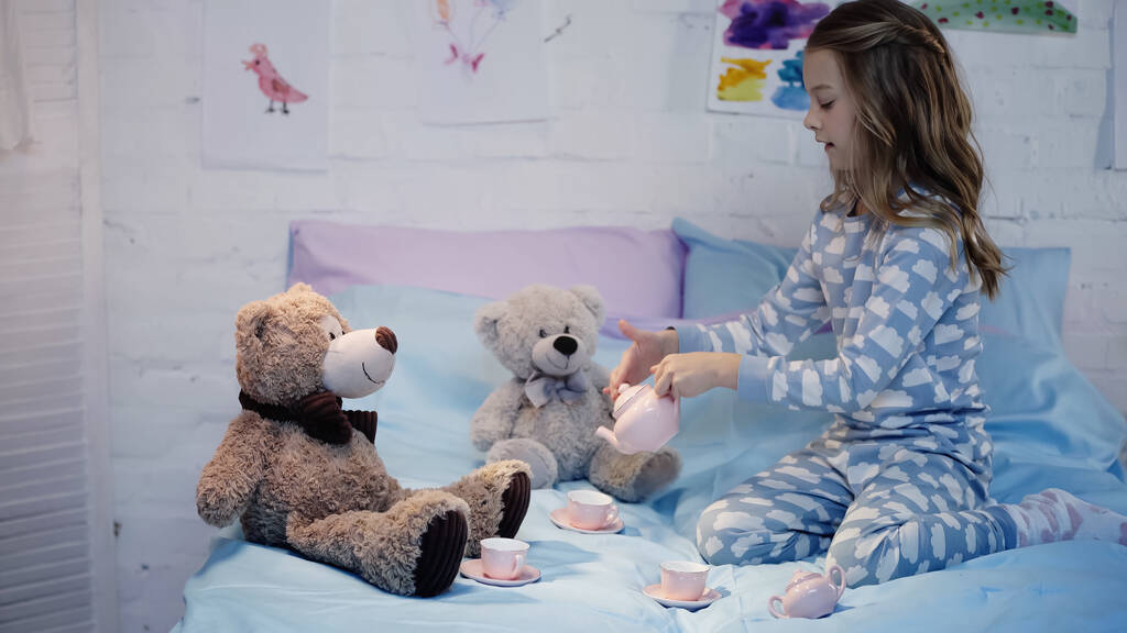 Side view of kid in pajama pouring tea near teddy bears on bed  - Fotó, kép