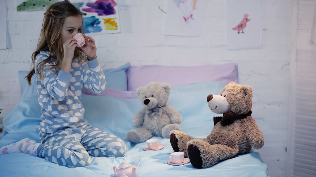 Preteen child drinking tea near soft toys on bed in evening  - Foto, immagini