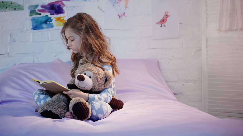 Pijama giymiş, kitap okuyan ve evdeki yatakta yumuşak oyuncaklar tutan bir çocuk.  - Fotoğraf, Görsel