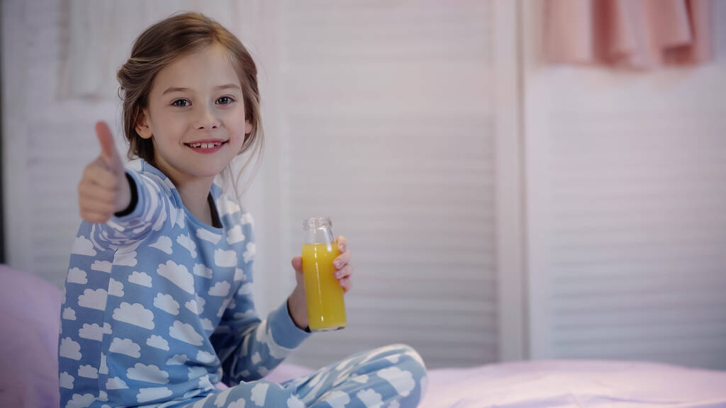 Pijamalı gülümseyen çocuk başparmağını kaldırıyor ve yatakta portakal suyu tutuyor.  - Fotoğraf, Görsel