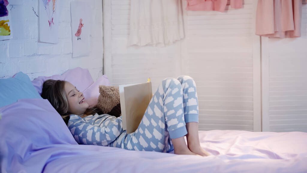 Preteen kid in pajama laughing while reading book near teddy bear on bed  - Foto, imagen