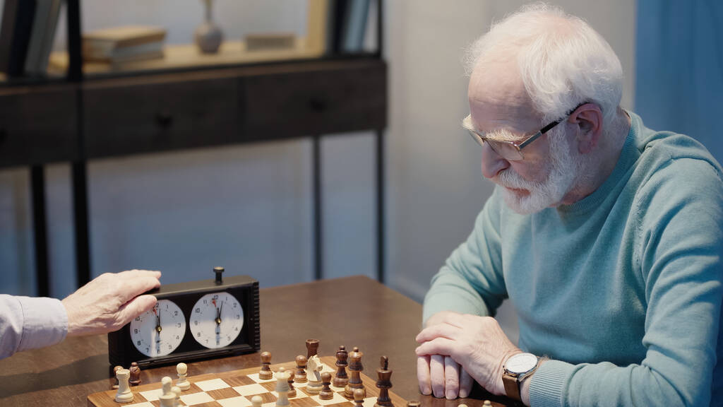 grey haired man thinking near chessboard and friend fixing time on chess clock - Foto, Imagen