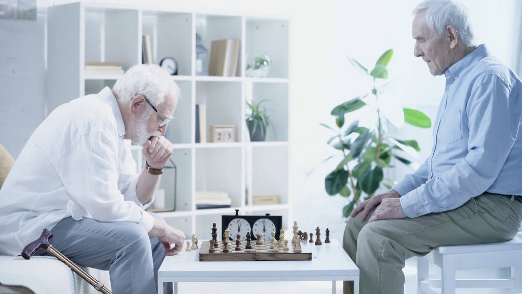 вид збоку вдумливого чоловіка, який грає в шахи з старшим другом у вітальні
 - Фото, зображення