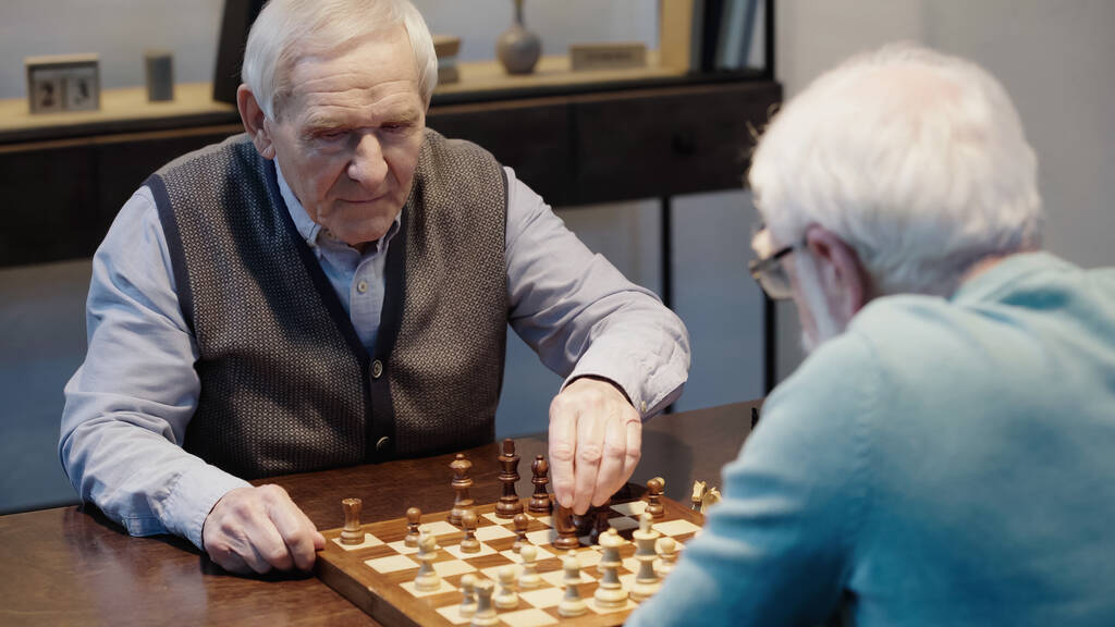grey haired man playing chess with blurred friend in living room - Fotografie, Obrázek