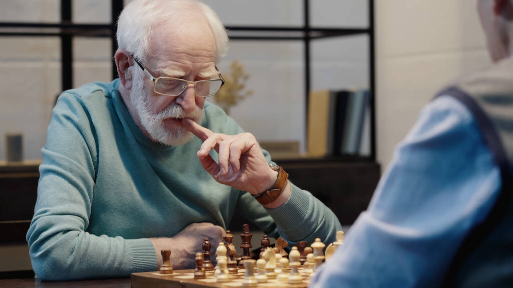 thoughtful senior man in eyeglasses touching lips while playing chess with blurred friend - Foto, afbeelding