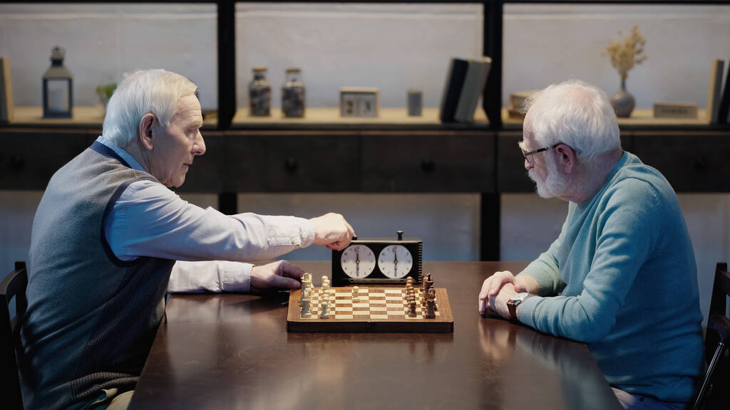 side view of grey haired man fixing time on chess clock near senior friend  - Foto, imagen