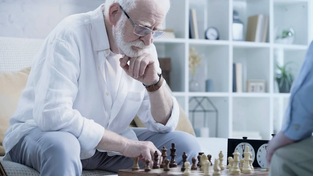 senior grey haired man in eyeglasses playing chess with friend in living room - Foto, afbeelding