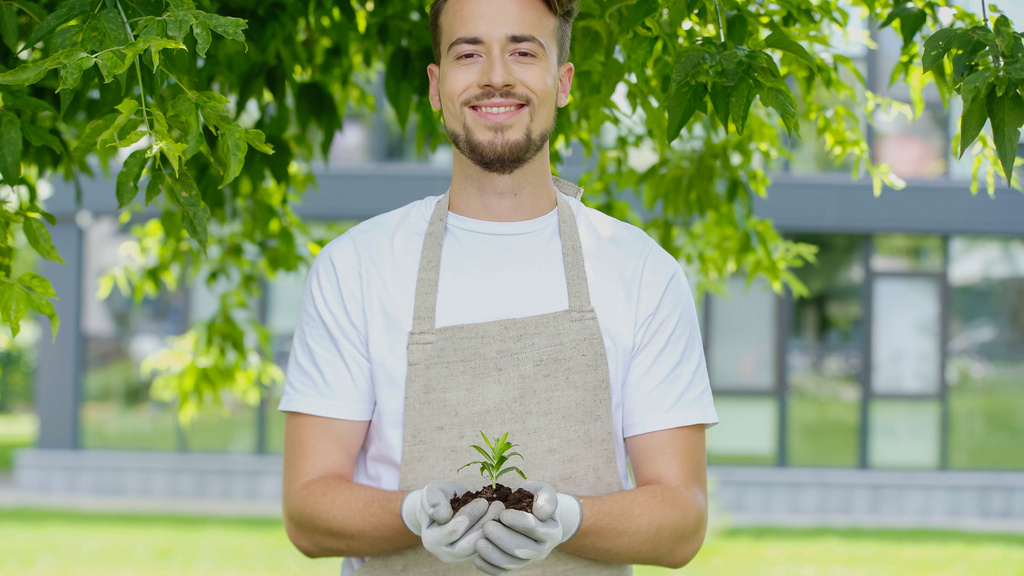 Smiling gardener in gloves holding plant in soil in garden  - Photo, image
