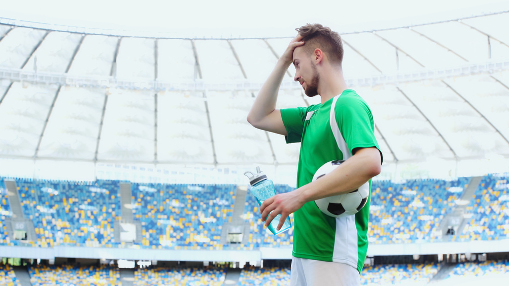 side view of young football player in green t-shirt holding ball while adjusting hair and holding sports bottle - Φωτογραφία, εικόνα