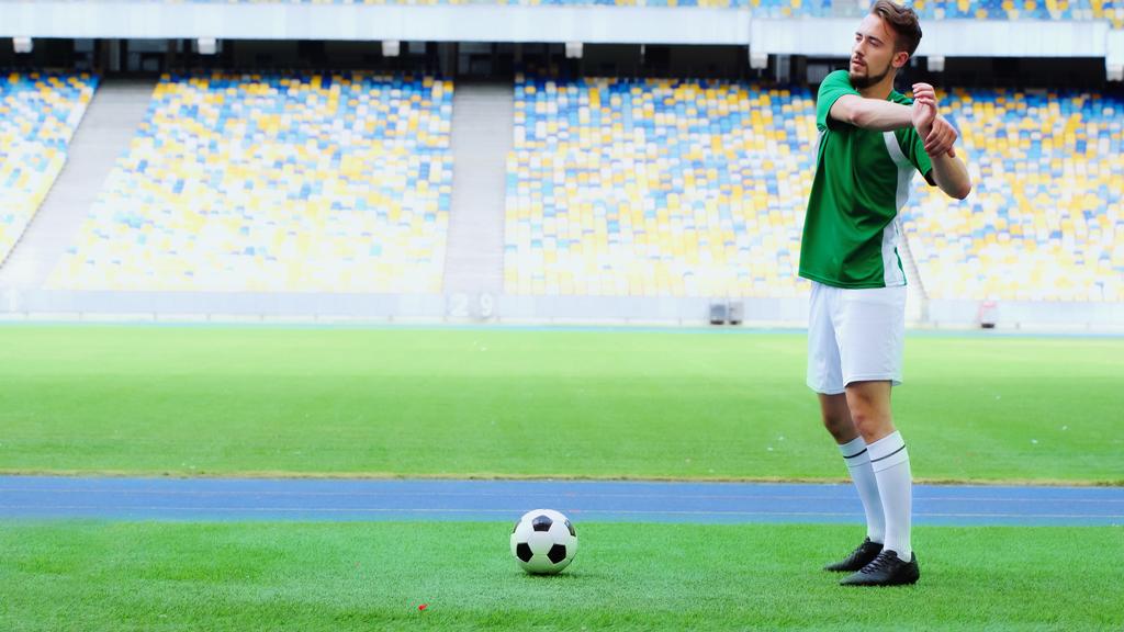full length of bearded football player in green uniform warming up near ball at stadium - Foto, afbeelding