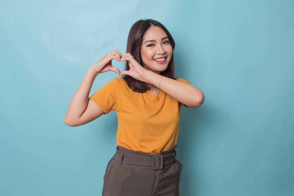 Attractive young Asian woman feels happy and romantic shapes heart gesture expresses tender feelings wears casual yellow t-shirt against blue background. People affection and care concept - Zdjęcie, obraz