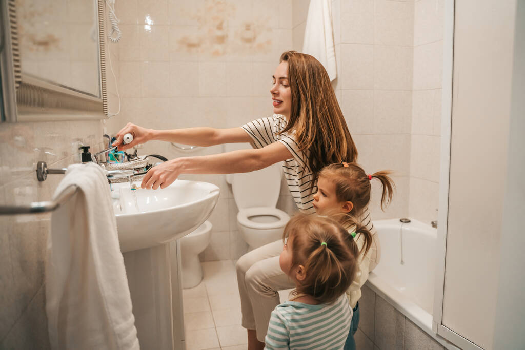 Усміхнена задоволена жінка змочує зубну щітку під проточною водою в присутності двох її дочок
 - Фото, зображення